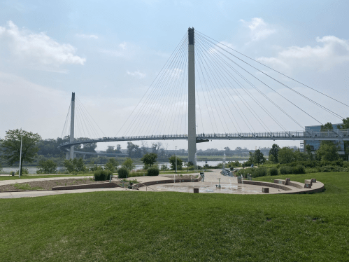 A modern cable-stayed bridge spans a river, surrounded by green grass and trees under a cloudy sky.