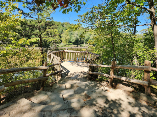 A wooden overlook surrounded by trees, leading to a scenic view of a river and lush greenery on a sunny day.