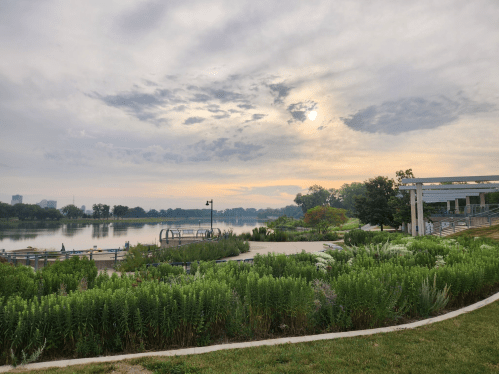 A serene riverside scene at dawn, featuring lush greenery, calm water, and a cloudy sky.