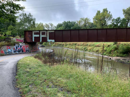 A rusted train bridge spans a river, with graffiti on the bridge and nearby walls, surrounded by greenery and a path.