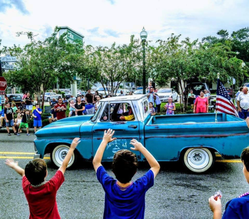 A blue vintage truck drives by a cheering crowd at a parade, with children waving and an American flag displayed.