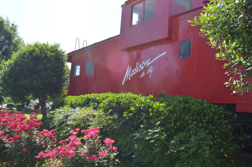 A bright red train caboose surrounded by green bushes and pink flowers in a sunny outdoor setting.