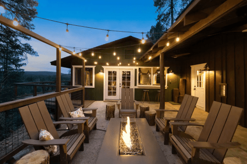 Cozy outdoor patio with string lights, wooden chairs, and a fire pit, surrounded by trees at dusk.