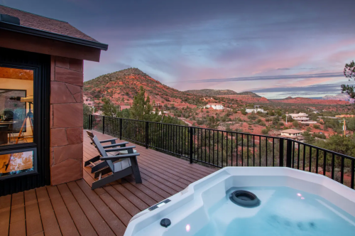 A hot tub on a deck overlooks a scenic view of mountains and a colorful sky at sunset.