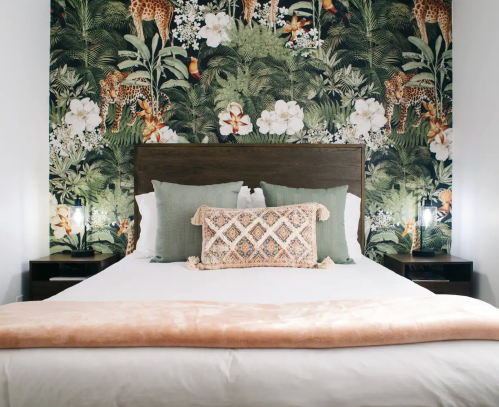 A stylish bedroom featuring a floral and jungle-themed wallpaper, a wooden bed frame, and decorative pillows.