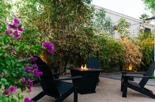 A cozy outdoor space with black chairs, a fire pit, and lush greenery, accented by blooming purple flowers.