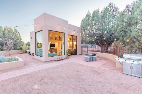 Modern small house with large windows, surrounded by trees and outdoor seating, featuring string lights and a grill.