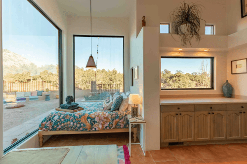 Cozy bedroom with large windows, colorful bedding, and a view of nature outside. Warm, inviting decor.