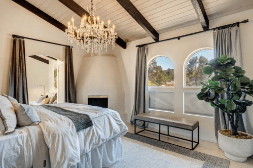 Bright bedroom with a chandelier, large windows, and a cozy bed, featuring modern decor and a potted plant.