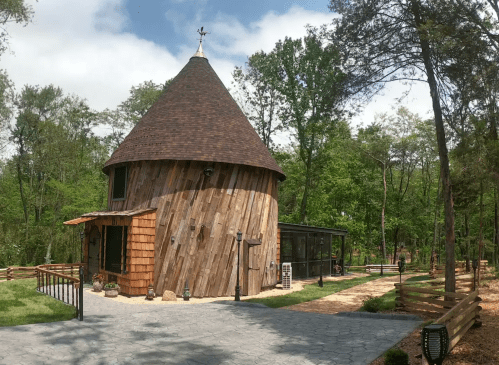 A unique, round wooden cabin with a conical roof, surrounded by trees and a stone pathway.