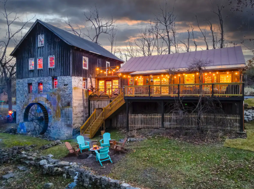 A charming rustic building with a water wheel, adorned with string lights, surrounded by a grassy area and chairs.