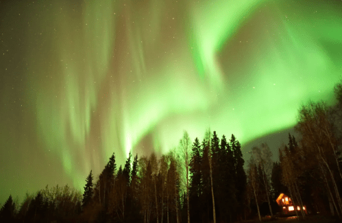 A stunning display of green auroras dances over a forest and a cozy cabin under a starry night sky.