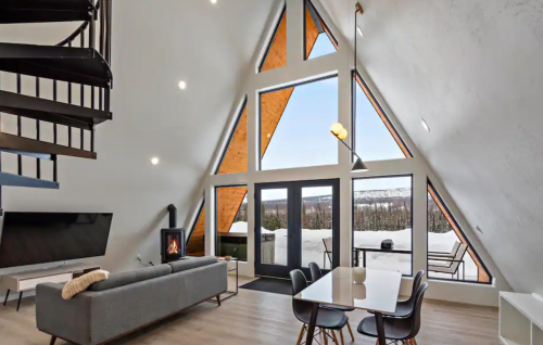 Modern A-frame cabin interior with large windows, a cozy fireplace, and a dining area, surrounded by snowy landscape.