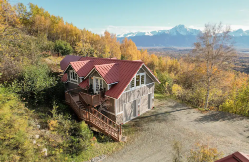 A cozy cabin with a red roof surrounded by autumn trees and mountains in the background.