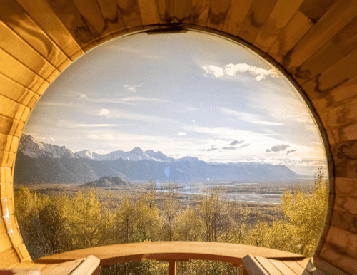 A circular window view of mountains and a valley, framed by wooden walls, with autumn foliage in the foreground.