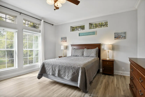 Bright bedroom with a queen bed, two lamps, and large windows, featuring light gray walls and wooden flooring.