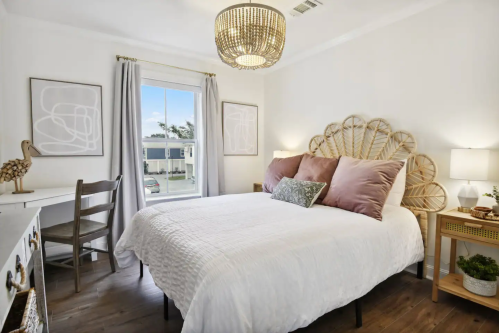 Cozy bedroom with a rattan headboard, white bedding, and natural light from a window, featuring a desk and decorative plants.