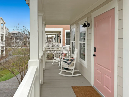 A porch with rocking chairs and a pink door, overlooking a neighborhood with houses and greenery.