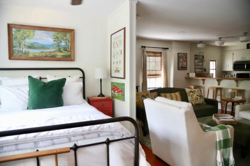 Cozy bedroom with a bed, green pillow, and artwork, leading to a living area with a sofa and kitchen in the background.