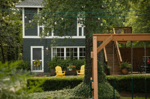 A charming two-story house with a gray exterior, yellow chairs on the porch, and a wooden swing set in the foreground.