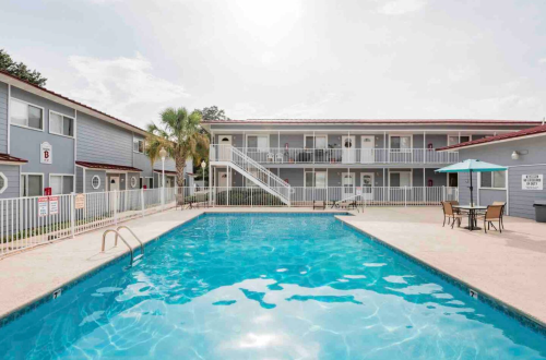 A clear blue pool surrounded by a two-story building with balconies and outdoor seating under a sunny sky.