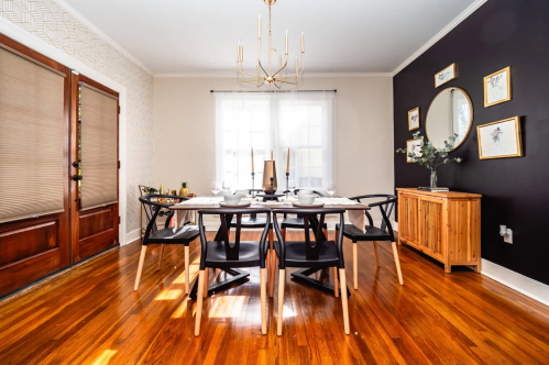 A modern dining room with a wooden table, black chairs, and a stylish chandelier, featuring large windows and decorative accents.