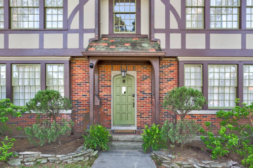 A charming brick and timber house with a green door, surrounded by shrubs and a stone pathway.