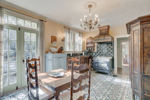 Bright kitchen with wooden furniture, patterned tile floor, and a vintage stove, featuring large windows and a chandelier.