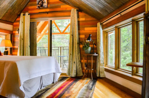 Cozy cabin interior with a bed, wooden walls, large windows, and a small table by the window, surrounded by nature.