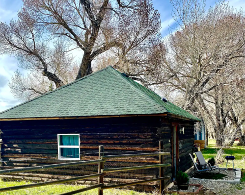 A rustic log cabin with a green roof, surrounded by bare trees and a wooden fence on a sunny day.