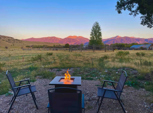 A cozy outdoor scene with a fire pit and chairs, overlooking mountains at sunset.