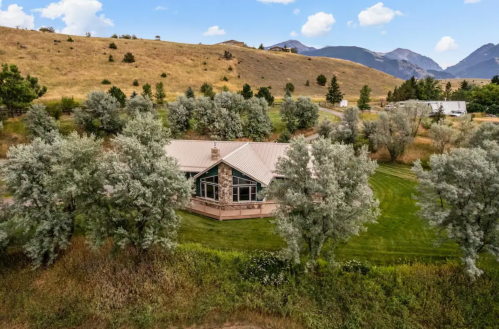 Aerial view of a house surrounded by trees and grassy land, with mountains in the background under a blue sky.
