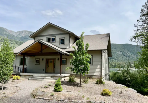 A modern two-story house surrounded by trees and mountains, with a porch and landscaped yard.
