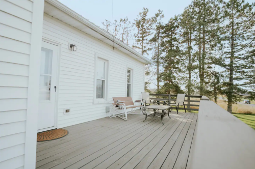 A wooden deck with seating, surrounded by trees and a clear sky, attached to a white house.