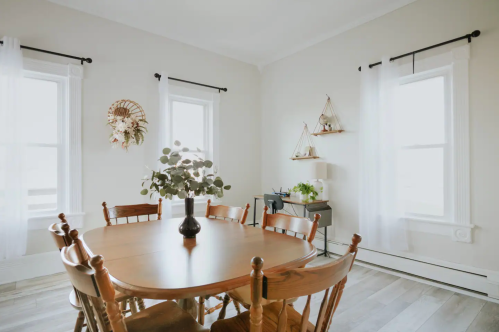 A bright dining room with a wooden table, chairs, and decorative plants near windows, creating a cozy atmosphere.
