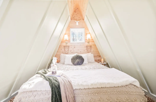 Cozy A-frame bedroom with a plush bed, soft blankets, and warm lighting, featuring wooden walls and a window.