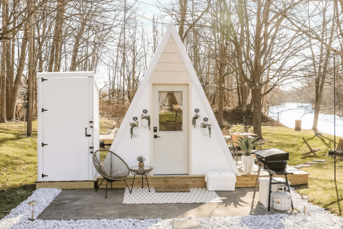 A modern A-frame cabin with a white exterior, surrounded by trees and a small patio area with outdoor furniture.