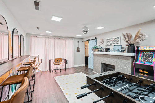 A stylish room with a foosball table, bar seating, desk, and arcade game, featuring large windows and modern decor.