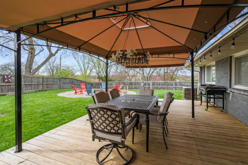 A covered patio with a dining table and chairs, overlooking a grassy yard with red chairs and a fire pit.