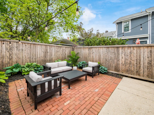 Cozy outdoor patio with seating, a coffee table, and greenery, enclosed by a wooden fence.
