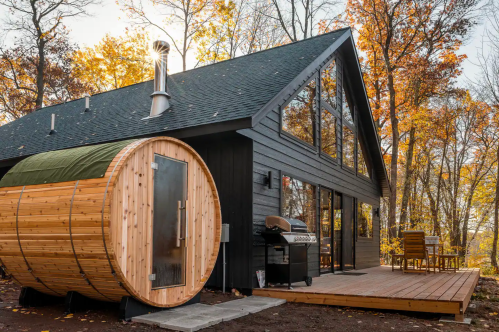 A modern cabin with large windows, a wooden sauna, and a grill on a deck surrounded by autumn trees.