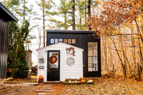 A charming tiny house with a white and black exterior, surrounded by autumn foliage and pumpkins.