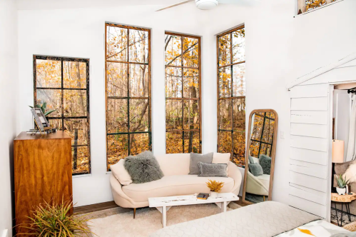 Cozy living space with large windows showcasing autumn trees, a sofa, mirror, and wooden dresser.