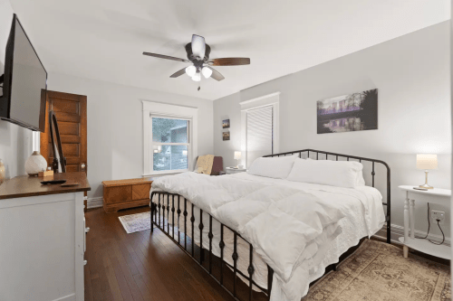 Cozy bedroom with a large bed, wooden furniture, a TV, and soft lighting, featuring a ceiling fan and neutral decor.