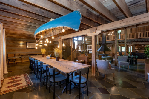 A rustic dining area with a long wooden table, blue canoe overhead, and warm lighting in a cozy, spacious setting.