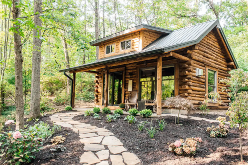 A charming log cabin surrounded by trees and landscaped gardens, featuring a stone pathway leading to the entrance.