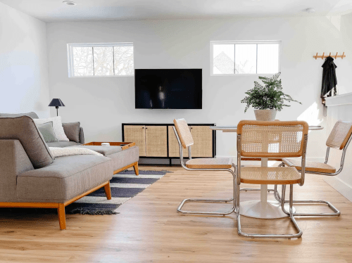 Modern living room with a gray sofa, TV, and dining table with chairs, featuring natural light and minimalist decor.