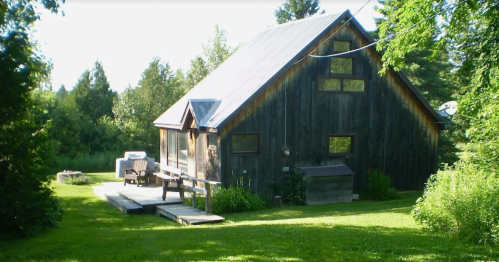 A rustic wooden cabin surrounded by greenery, featuring a deck and outdoor seating area in a sunny landscape.
