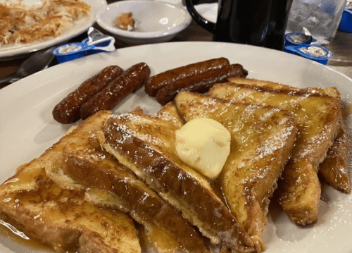 A plate of French toast topped with butter and syrup, accompanied by sausage links and a coffee mug in the background.