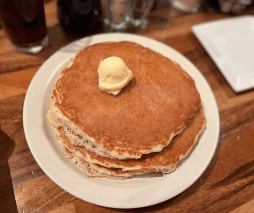 A stack of three golden-brown pancakes topped with a pat of butter on a white plate, set on a wooden table.